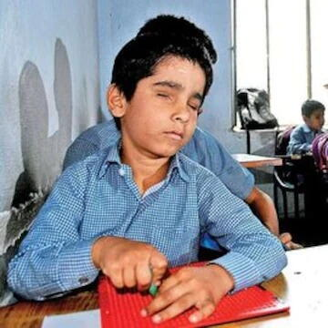 A visually impaired student writing Braille with a stylus and slate at his classroom desk
