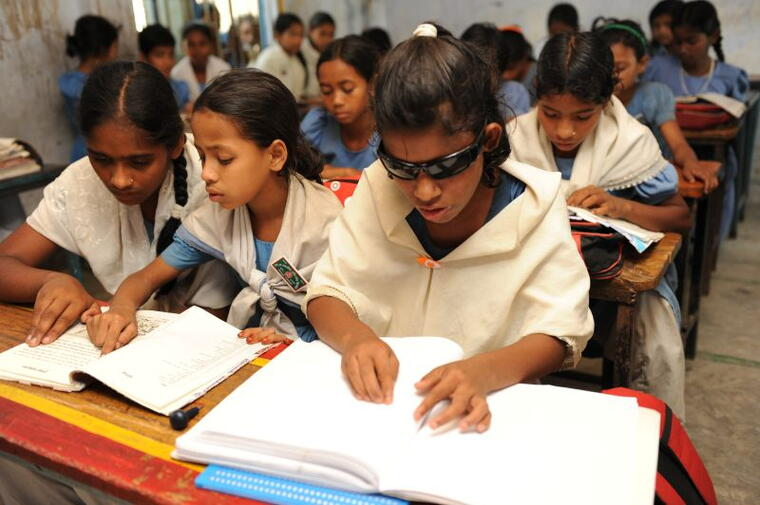 Girls reading Braille books together in a classroom, one wearing protective tinted glasses